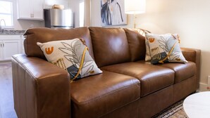 Un canapé en cuir marron agrémenté de deux coussins décoratifs, une cuisine en arrière-plan et un tableau accroché au mur.
