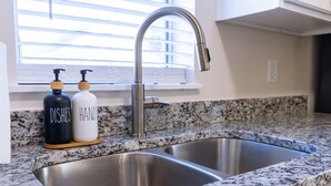 Une cuisine équipée d’un évier en acier inoxydable à deux bacs, d’un plan de travail en granit et de deux distributeurs de savon, l’un portant l’inscription « VAISSELLE » et l’autre « MAINS ».