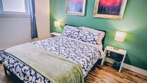 Un dormitorio con cama, dos mesitas de noche, un ventanal con cortinas y dos cuadros colgados en la pared.
