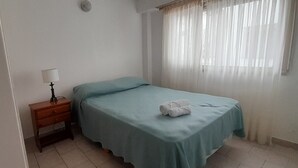 Een enkel bed met een blauw sprei, een houten nachtkastje met een lamp, en een raam met transparante gordijnen.