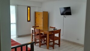 Een kamer met een houten eettafel, vier stoelen, een aan de muur bevestigde televisie en een raam met jaloezieën.