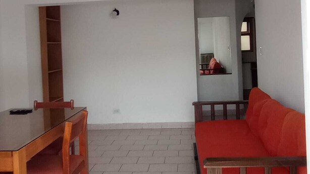Een woonkamer met een eettafel en stoelen, een rode bank, een houten boekenkast en een betegelde vloer.