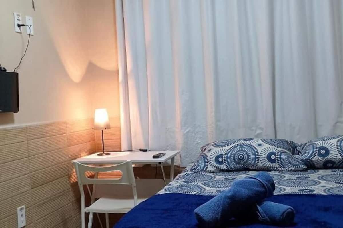 A hotel room with a bed, a desk with a lamp, a chair, and a wall-mounted TV.