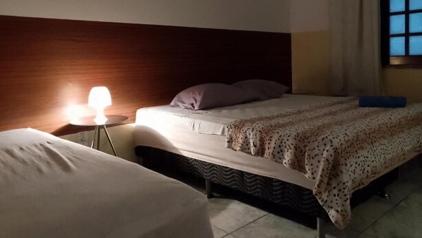 A hotel room with a bed, bedside table, lamp, and a window. - Hostel Aldeia Inn (Fortaleza)