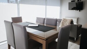 A modern dining area with a glass table, cushioned chairs, a sofa, and a mounted TV.