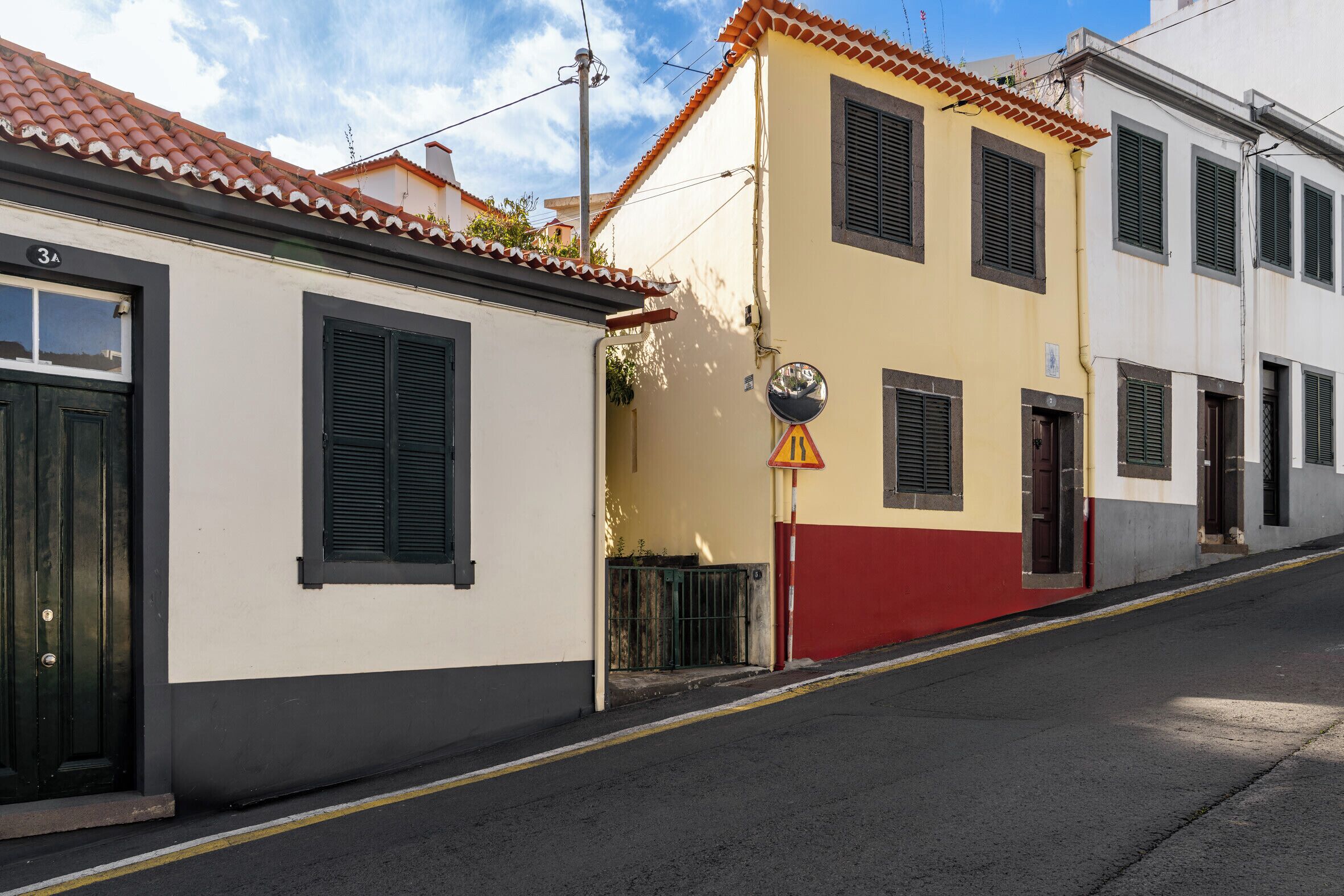 Eine Straße mit Gebäuden, darunter ein Haus mit der Nummer 3A an der Tür, ein gelb-rotes Gebäude und ein weißes Gebäude mit Fensterläden.