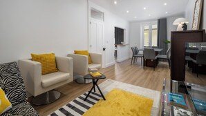 Una sala de estar moderna con un sofá, un sillón y una mesita con libros. Se observa un comedor con sillas y una mesa al fondo.