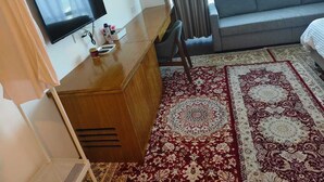 Une chambre d’hôtel avec un bureau en bois, un téléviseur à écran plat fixé au mur, un canapé gris et un tapis rouge et blanc à motifs.