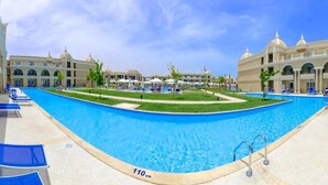 A swimming pool with a 110 cm depth, surrounded by lounge chairs and umbrellas, with a large building in the background. - Titanic Royal Palace (Hurghada)