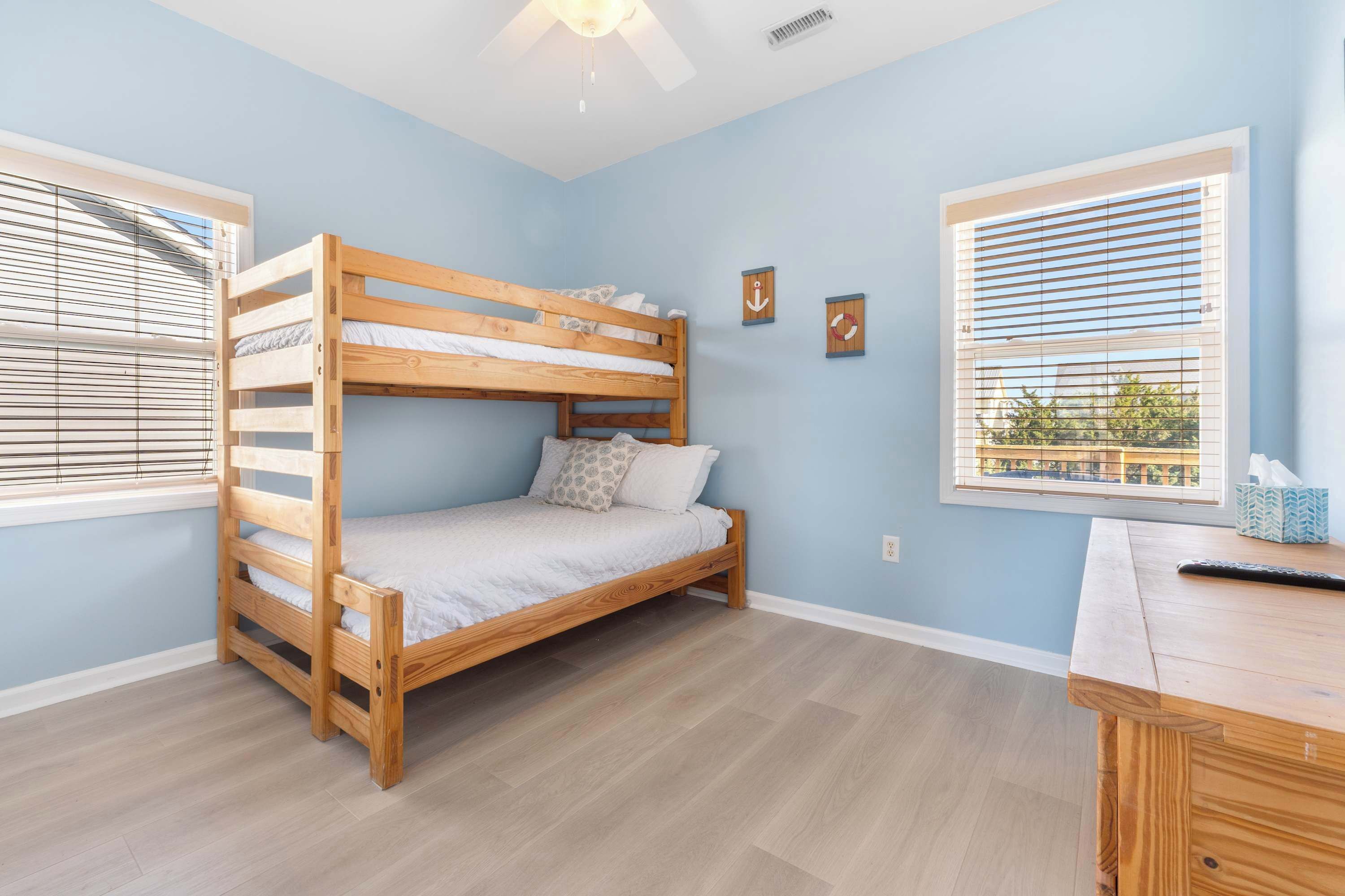 A bunk bed in a room with light blue walls, a wooden desk, and two windows with blinds.