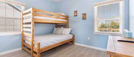 A bunk bed in a room with light blue walls, a wooden desk, and two windows with blinds.