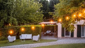 Een terras buiten met een pergola, lichtslingers en een haag.