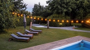 Een achtertuin met een zwembad, lounge stoelen, een hangmat, lichtjes en een houten terras.