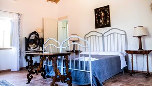 Een slaapkamer met een bed, een tafel, een stoel, een lamp, een spiegel en een schilderij aan de muur.