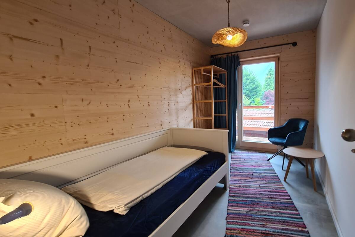 Une pièce comprenant un lit, une chaise, une petite table, un luminaire suspendu et une fenêtre donnant sur un paysage verdoyant.