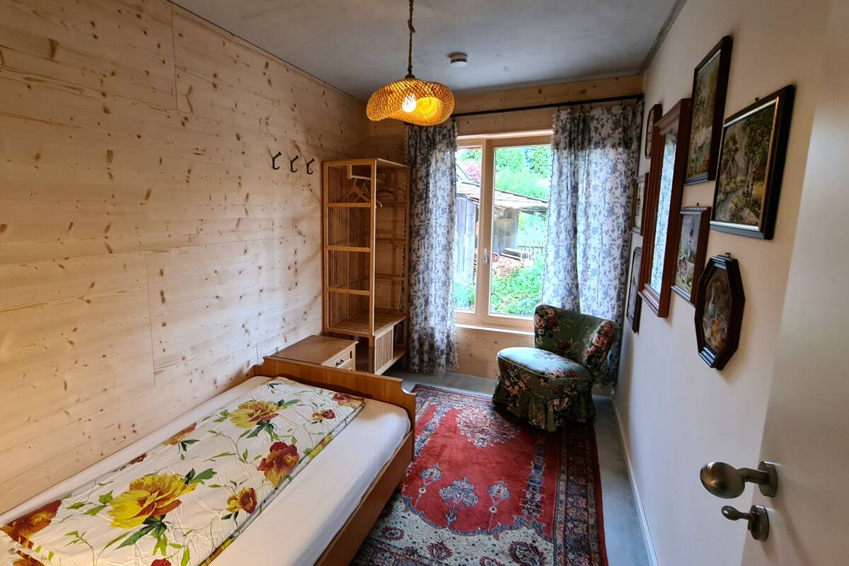 Une pièce avec un mur en bois, un lit recouvert d’une literie à motifs floraux, une chaise, un luminaire suspendu et une porte dotée d’une poignée.
