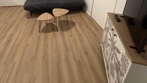 Een kamer met een donkergrijze bank, twee kleine houten tafels, een televisie op een houten dressoir en een deur.
