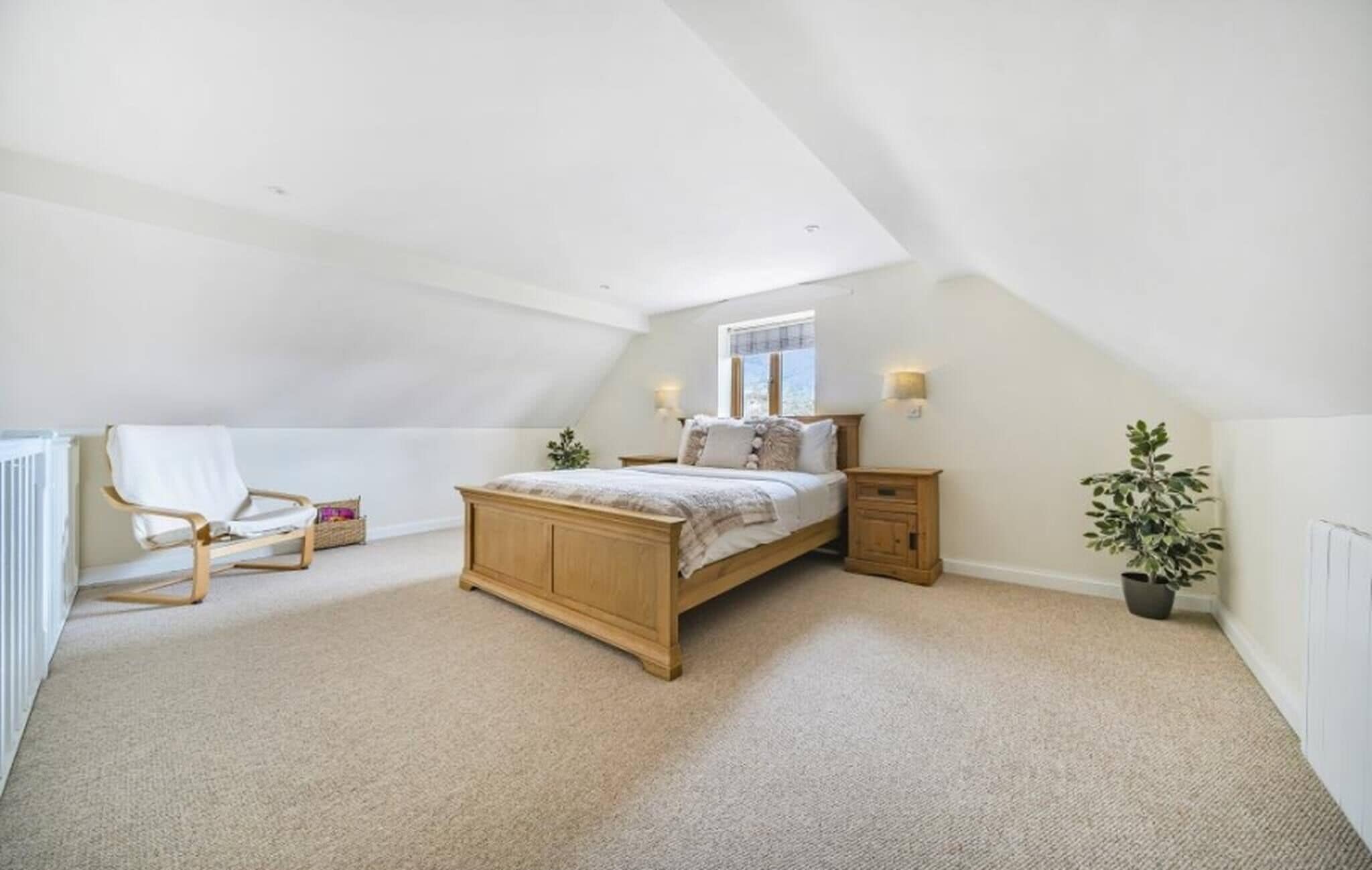 A bedroom with a sloped ceiling, a bed, a chair, bedside tables, and a potted plant.