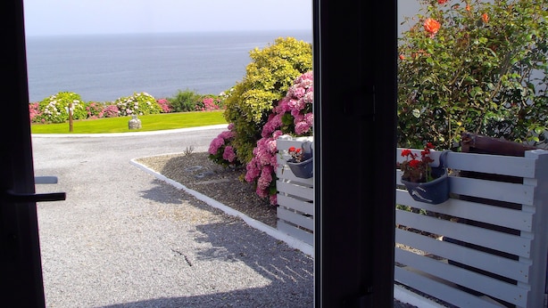 Une vue à travers l’encadrement d’une porte donnant sur un jardin, avec un chemin de gravier menant à une clôture blanche, un banc et diverses plantes en fleurs.