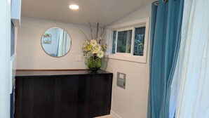 A bathroom with a dark wooden vanity, a round mirror, a vase with flowers, a window with blue curtains, and a wall-mounted electrical outlet.