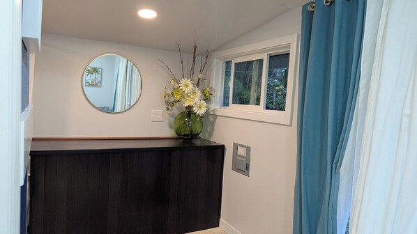 Une salle de bain avec un meuble-lavabo en bois foncé, un miroir rond, un vase avec des fleurs, une fenêtre avec des rideaux bleus et une prise électrique murale.