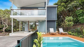 Una casa moderna de dos pisos con balcón, piscina y abundante vegetación.
