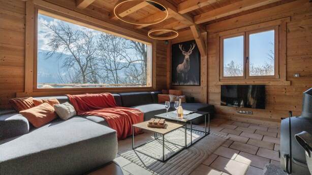 A log cabin interior with a sofa, fireplace, large windows, and a ceiling light fixture.