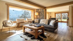 A cozy living room with a stone wall, wooden beams, a large window, a leather rug, and a sofa with pillows.