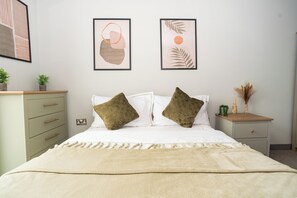 Een keurig opgeruimde slaapkamer met een bed, een nachtkastje, een kaptafel en een dressoir, en twee ingelijste kunstwerken aan de muur.