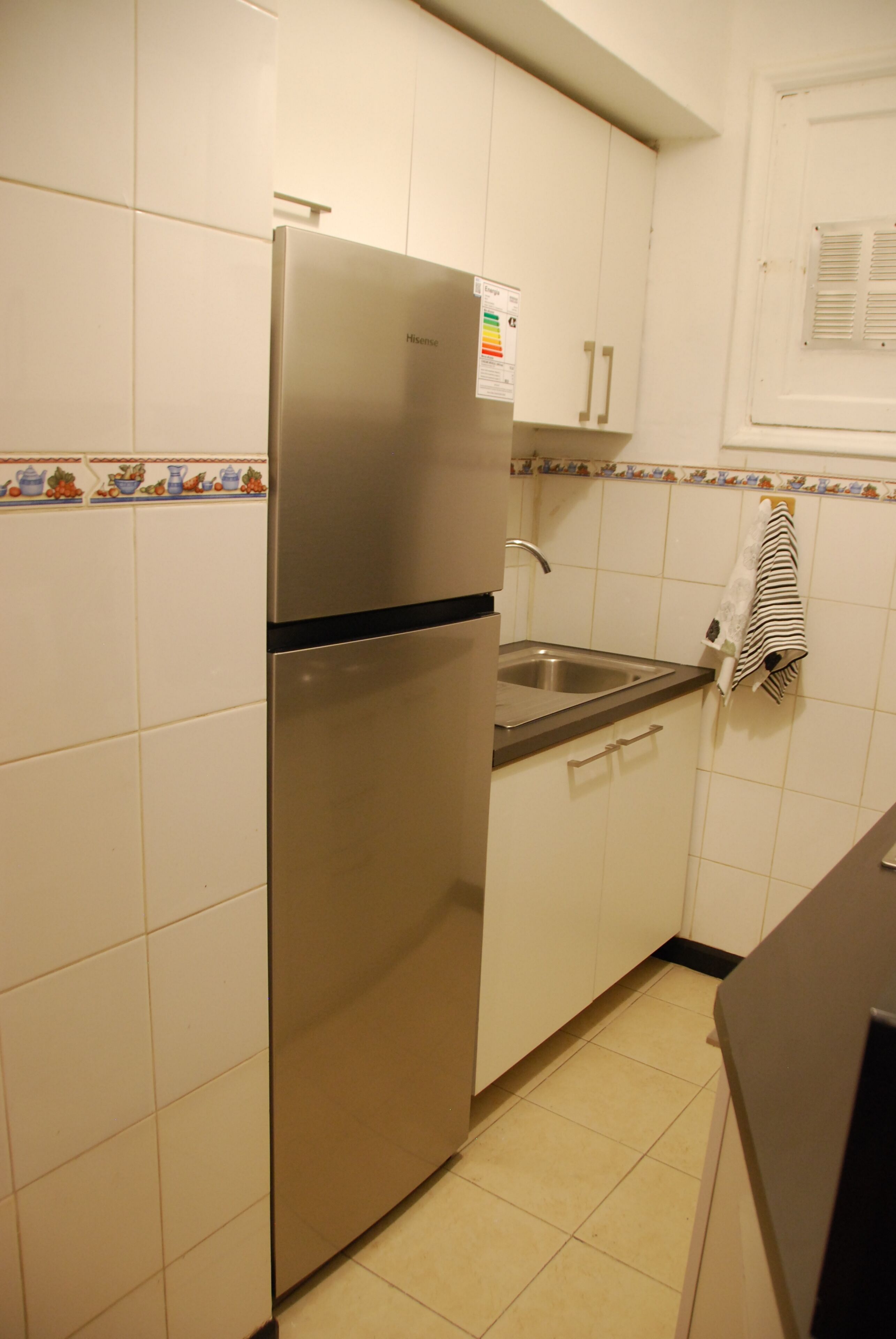 A kitchen with white cabinets, a stainless steel refrigerator, a sink, and a window with blinds.