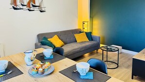 Ett modernt vardagsrum med en grå soffa, gula kuddar, ett matbord med en blå bordsduk och en väggdekoration i form av en segelbåt.