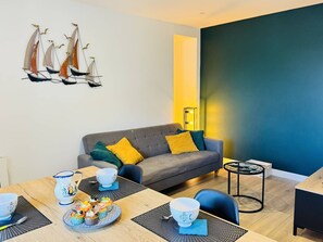 A modern living room with a grey sofa, yellow cushions, a dining table with a blue placemat, and a wall-mounted sailboat decoration. - 3 pièces plein centre ville, wifi, 5 min à pied plage (QUIBERON)