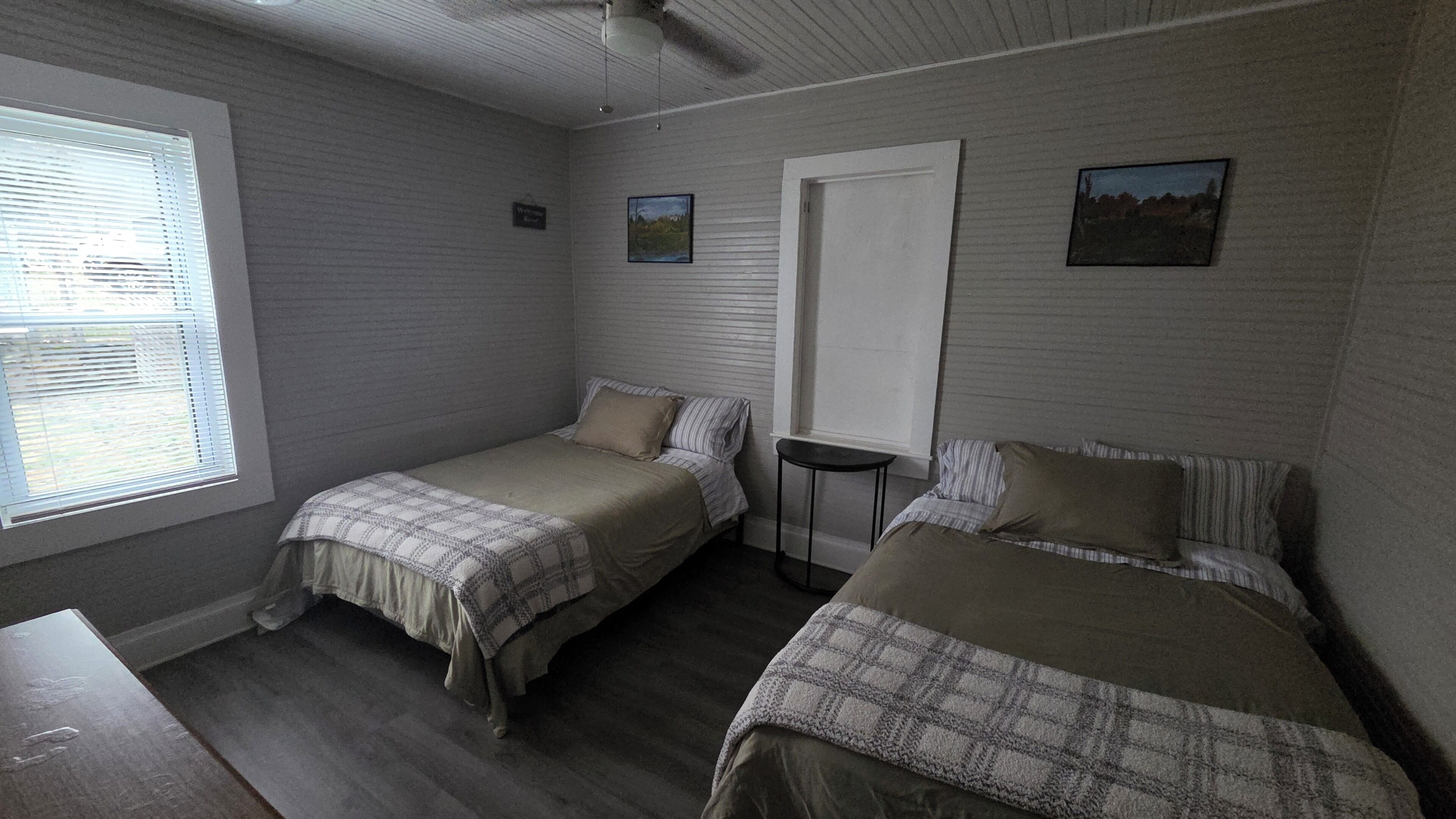 A bedroom with two single beds, a small table, a door, and a window with blinds.