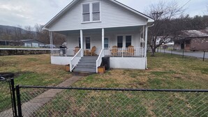 A two-story house with a covered porch, a staircase leading to the entrance, and a fenced yard. - New River Gorge Riverview Home in Meadow Creek.  3 bedroom, 1 bath, fenced yard (Meadow Creek)