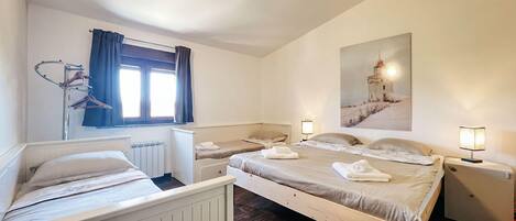 Quarto com uma cama grande, duas camas menores, um radiador, janela com cortinas, um quadro e luminárias de cabeceira.