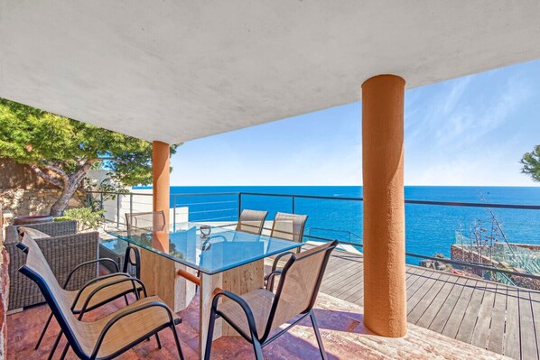 A glass table with chairs on a wooden deck overlooking the sea.