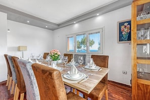 A dining area with a wooden table set for six, surrounded by upholstered chairs, a vase of flowers, and a view of the sea through large windows.