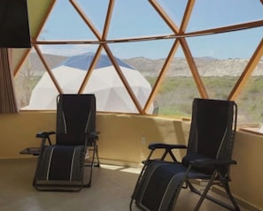A room with two reclining chairs, a large window with a geometric frame, and a view of a mountainous landscape. - Glamping Uno los Barilles (Los Barriles)
