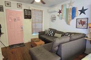 Ein Wohnzimmer mit grauem Sofa, einer pinkfarbenen Tür, einem Fenster mit Jalousien und Wanddekorationen, darunter Sterne und ein Schild mit der Aufschrift Texas.