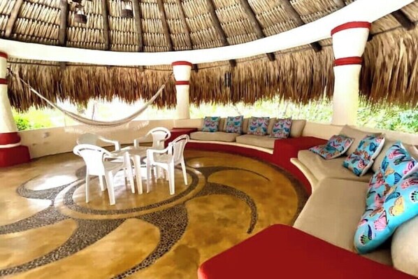 A circular seating area with a thatched roof, white chairs, and colorful cushions.