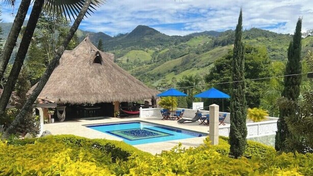 Zona de piscina con una estructura de techo de paja, sillas de descanso y vistas a colinas verdes.