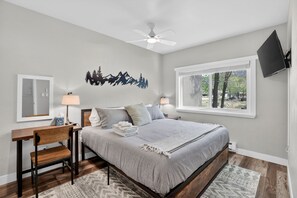 A bedroom with a bed, bedside tables, a desk, a chair, a ceiling fan, a TV, and a window with a view of trees.