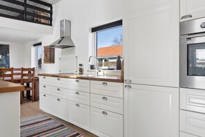 A modern kitchen with white cabinetry, a stainless steel oven, and a window providing a view of the outside. - Lovely holiday home near Hasmark Beach (Otterup)