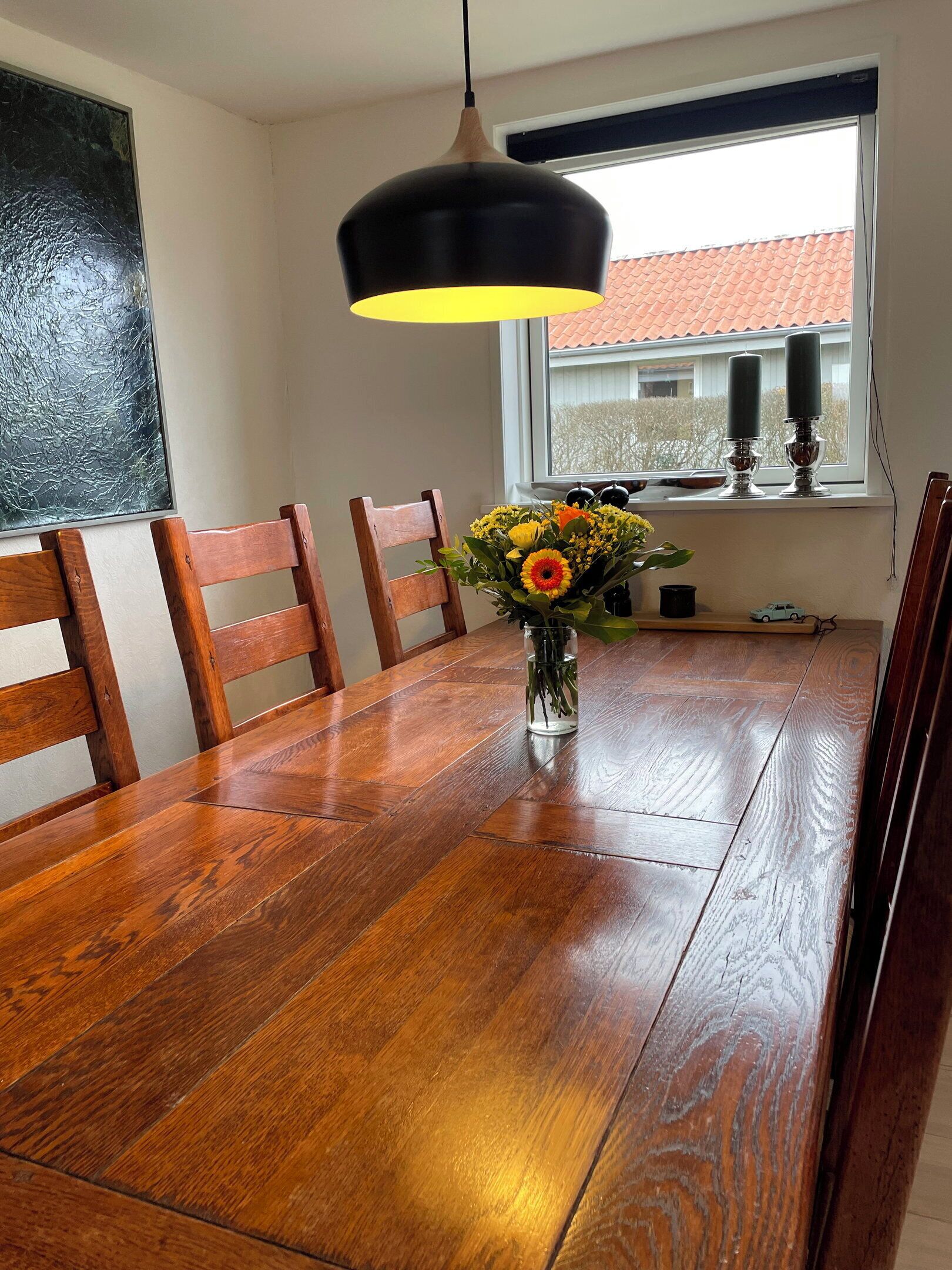 A dining table with chairs, a vase with flowers, a hanging light fixture, and a window with a view of a building.