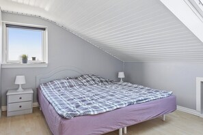 A bedroom with a sloped ceiling, a bed with a checkered blanket, a nightstand with a lamp, and a window with a view. - Lovely holiday home near Hasmark Beach (Otterup)