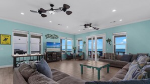 A spacious living room with a large sofa, a coffee table, and a TV. There are ceiling fans, a large window with a view, and a door leading to another room. - Baywatch Beach House | Private Pool | Beach Service Included | Dog Friendly (Pensacola Beach)