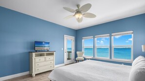 A bedroom with a bed, a dresser, a chair, a ceiling fan, and a view of the ocean through large windows. - Baywatch Beach House | Private Pool | Beach Service Included | Dog Friendly (Pensacola Beach)