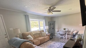 A living room with a sofa, a dining table with chairs, a ceiling fan, and a television.