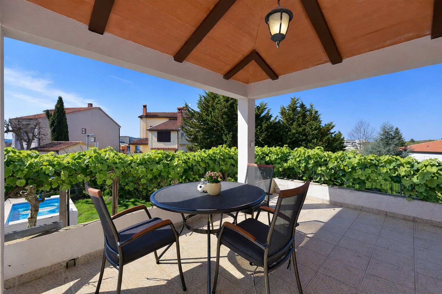 Eine Außenpatio mit einem runden Tisch und Stühlen, ein Pool im Hintergrund sowie Blick auf Häuser und Grünflächen.