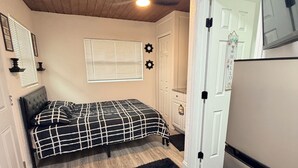 Une chambre avec un lit, un tapis noir, un ventilateur de plafond, une fenêtre avec des stores et une porte sur laquelle est inscrit le mot « BONJOUR ».